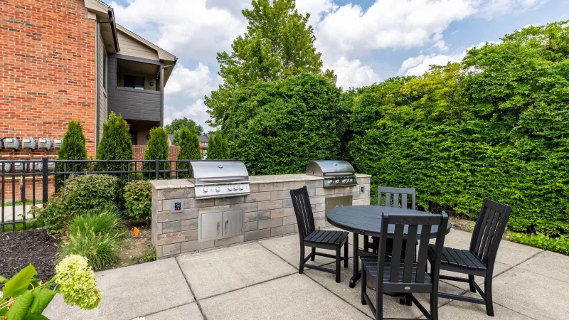 Outdoor patio with two built-in grills, a black dining set, and lush greenery next to a brick building.