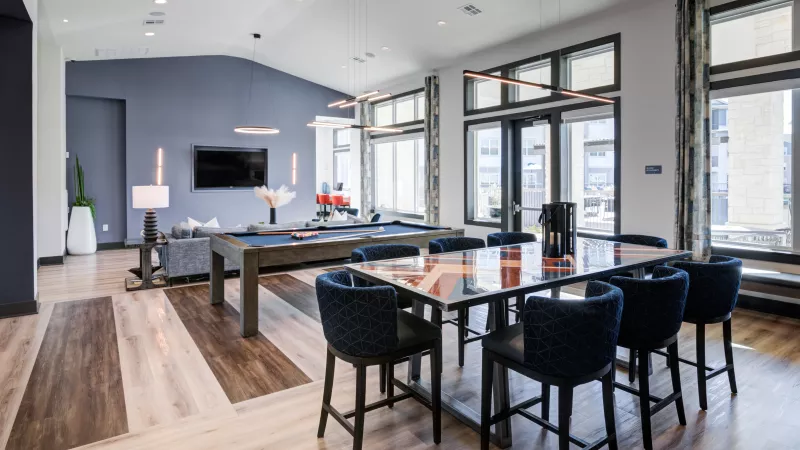 Spacious modern lounge featuring a pool table, dining area with blue chairs, and a TV lounge. High ceilings, large windows.