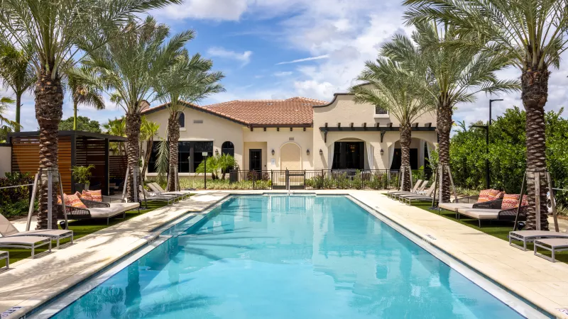 Luxury community pool with clear blue water, lined by palm trees and lounge chairs, in front of a light-colored building.