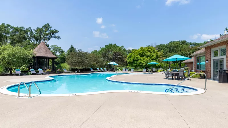 Sunny outdoor swimming pool with lounge chairs, a gazebo, and a brick clubhouse with outdoor seating.