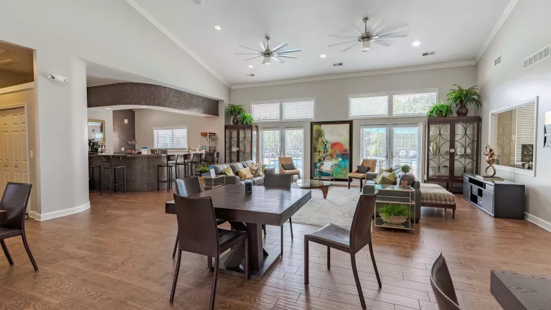 Spacious common room with dark dining table, lounge seating, and a bar area, bright and open with large windows.