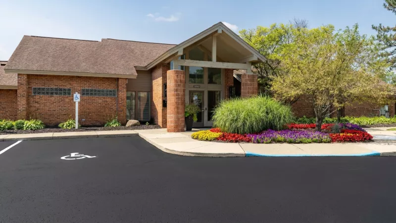 Photograph of a brick building entrance with a peaked roof, colorful flower beds, and green landscaping on a sunny day.