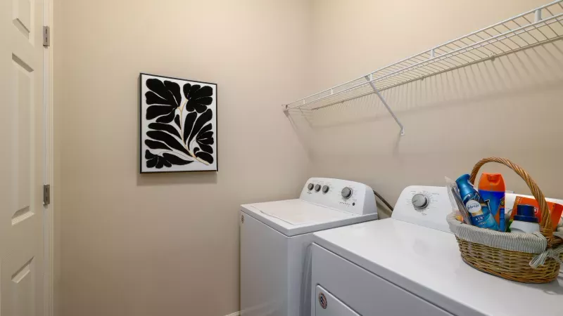 Beige laundry room with white washer, dryer, wire shelf, a basket of detergents, and black and white abstract art.