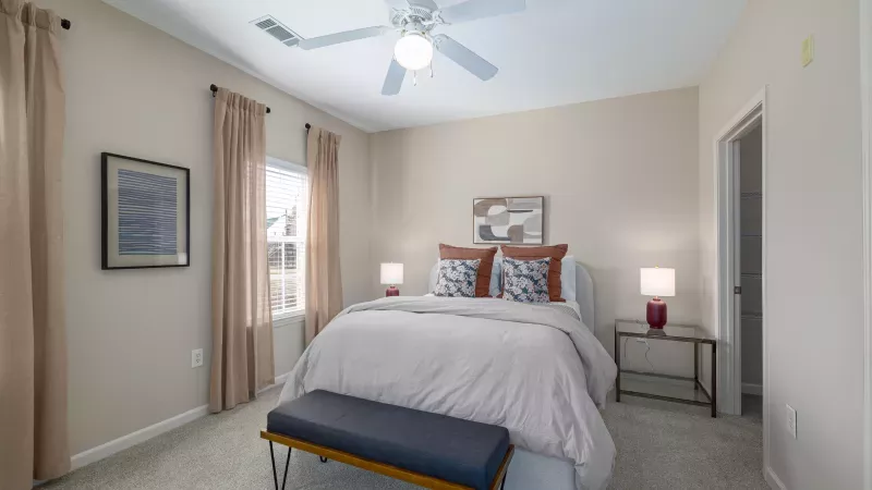 Bright bedroom with queen bed, dark blue bench, beige walls, light curtains, two nightstand lamps, and a ceiling fan.