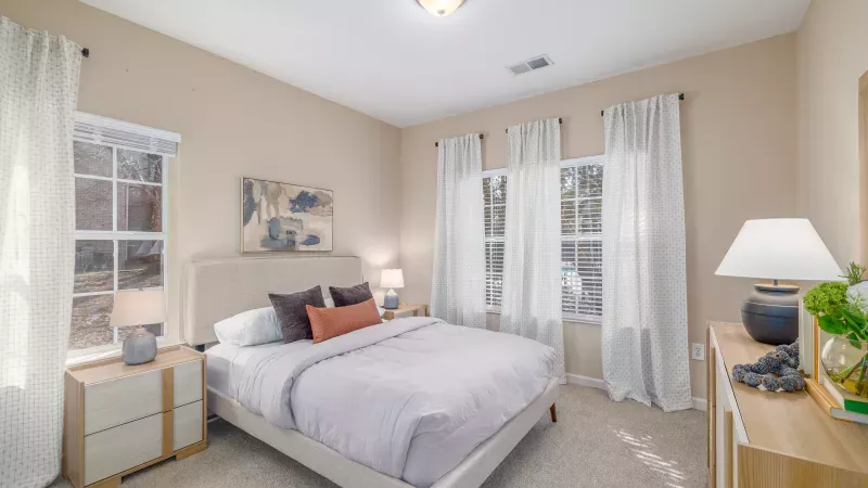 A bright bedroom with a bed, two nightstands, lamps, a dresser, and several windows with white sheer curtains.