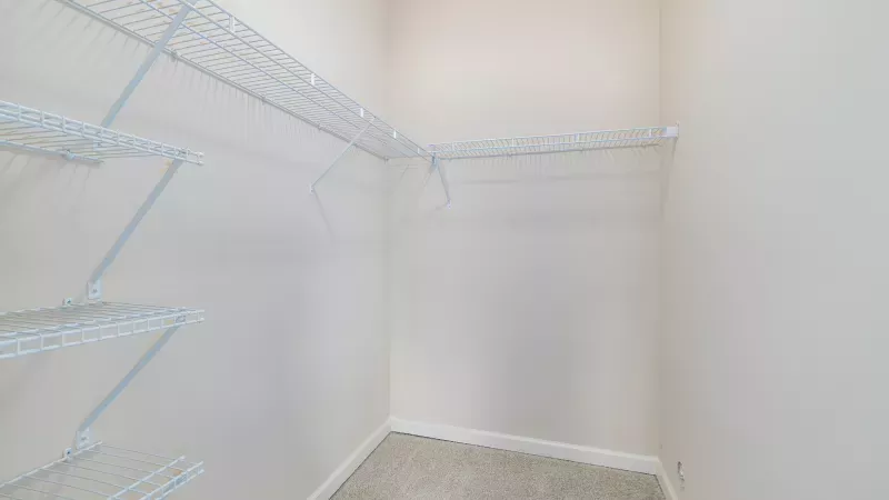 Empty walk-in closet with white wire shelving on three walls, light beige walls, and gray carpet.