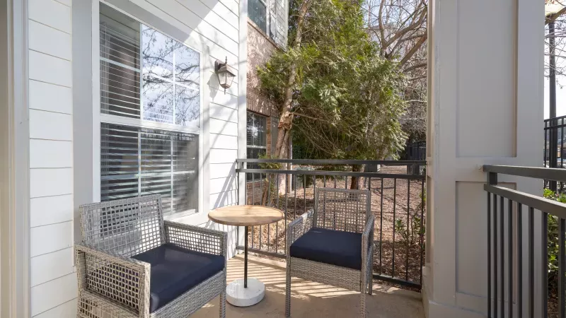 Sunny balcony with two grey woven chairs, dark cushions, and a small round table. Black railing and trees visible.