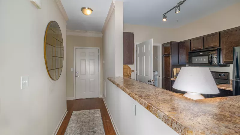 Hallway with a round mirror, white door, and a patterned rug leading to a kitchen with a speckled counter and dark cabinets.