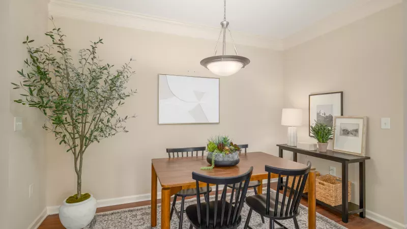 A modern dining room features a wooden table with black chairs, a chandelier, a large potted plant, and wall art.