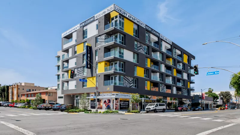 Photograph: Modern building with dark grey, yellow, white, and light grey panels and balconies, at a busy city street corner.