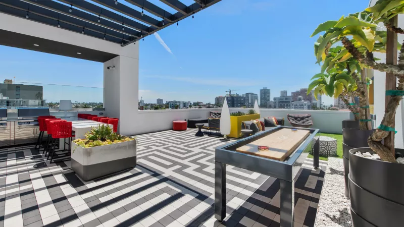 Modern rooftop patio with black and white patterned tiles, red bar stools, colorful lounge seating, a shuffleboard table, and city views.