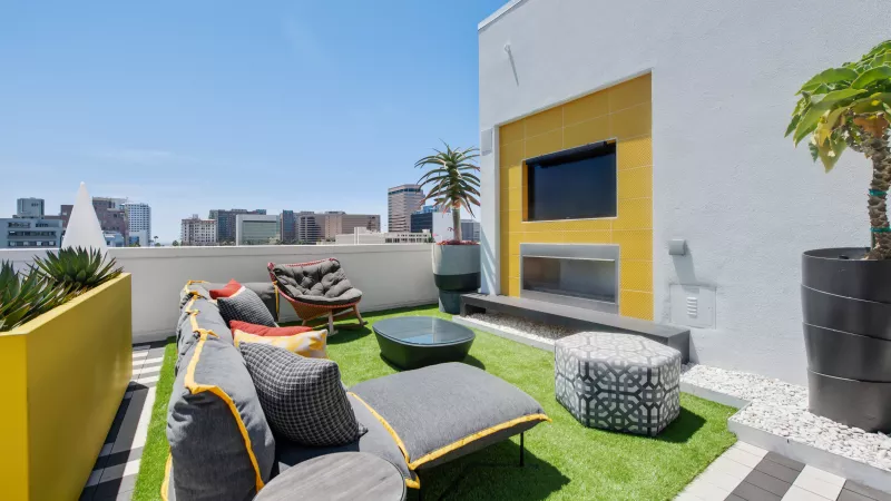 Modern rooftop patio with a grey sofa, green turf, a yellow accent wall with TV, potted plants, and a city skyline.