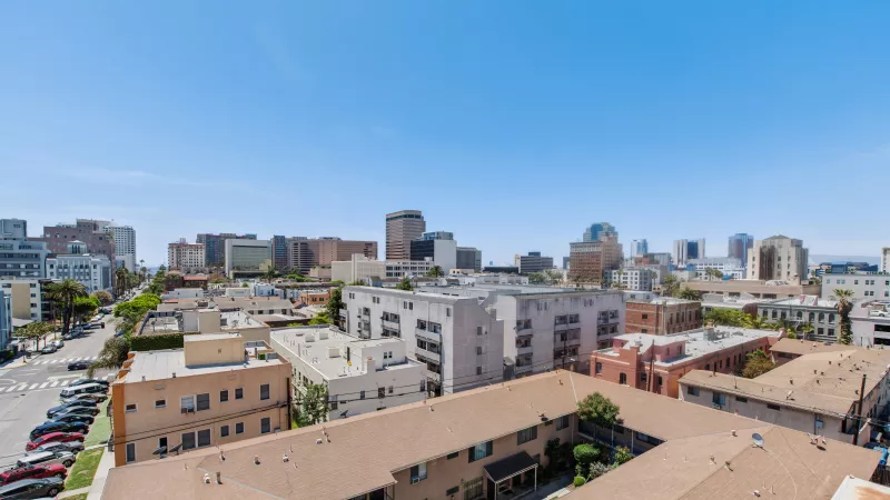 Aerial photograph of an urban landscape showing various buildings, a street, and a distant city skyline under a clear blue sky.