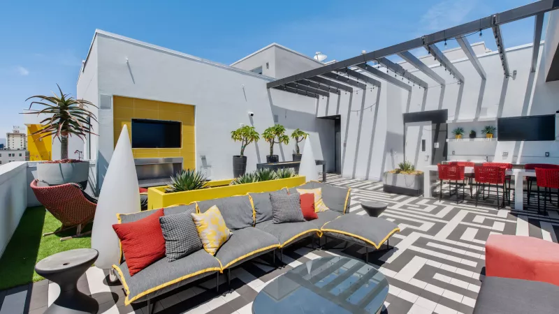 Modern rooftop patio with gray sectional, red chairs, yellow accents, and black and white geometric flooring under a clear sky.