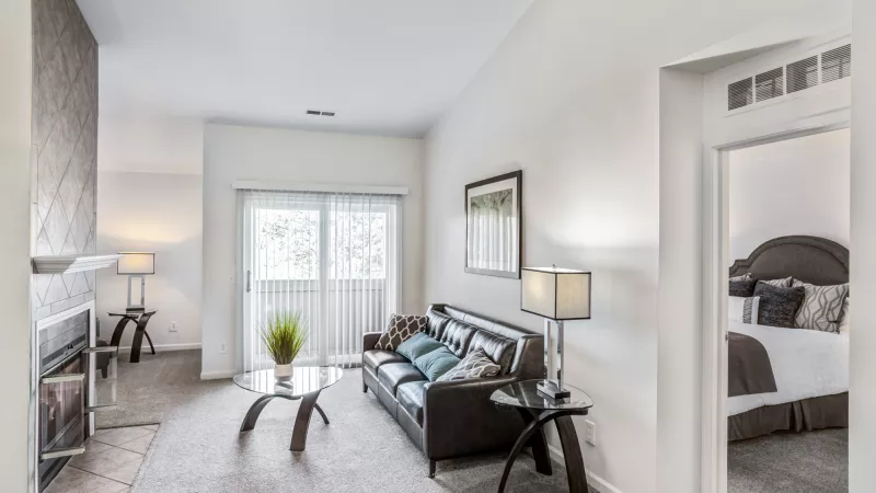Modern living room with dark leather sofa, glass table, fireplace, and sliding door. Bedroom visible through a doorway.