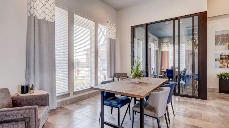 Modern dining area with a wooden table, blue and gray chairs, large windows with curtains, and a glass partition.