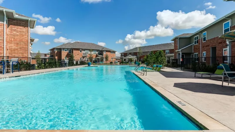 Large, bright blue swimming pool with lounge chairs, surrounded by brick apartment buildings under a sunny sky.