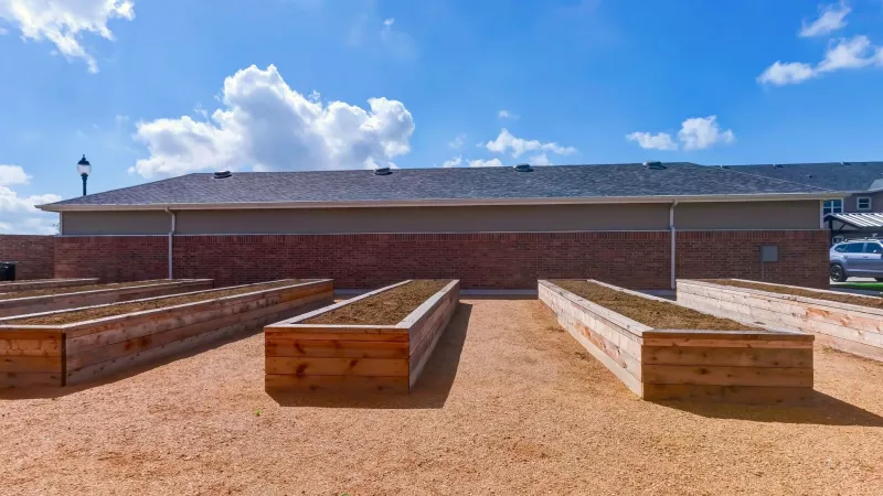 Raised wooden garden beds filled with soil on gravel, with a brick building and blue sky in the background.