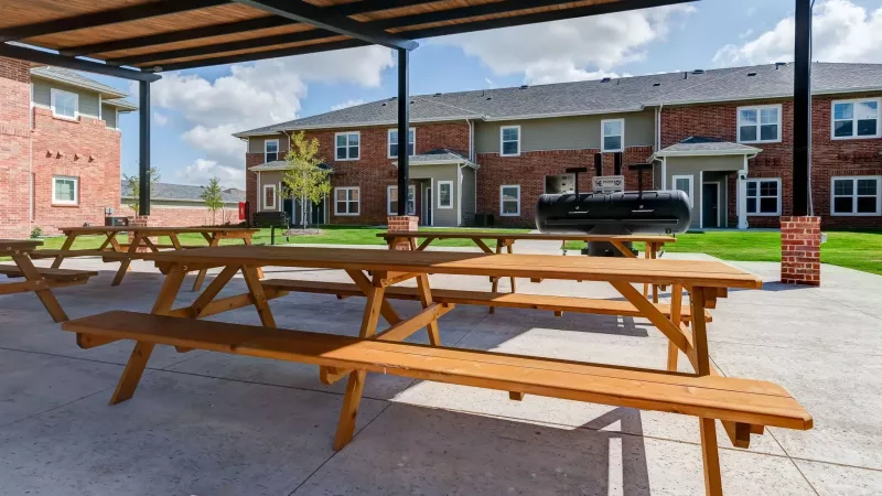 Outdoor patio area with several wooden picnic tables under a shaded pergola, a green lawn, and apartment buildings behind.