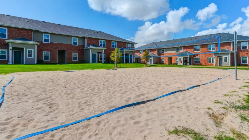 Sand volleyball court with blue lines in front of two apartment buildings under a sunny sky.