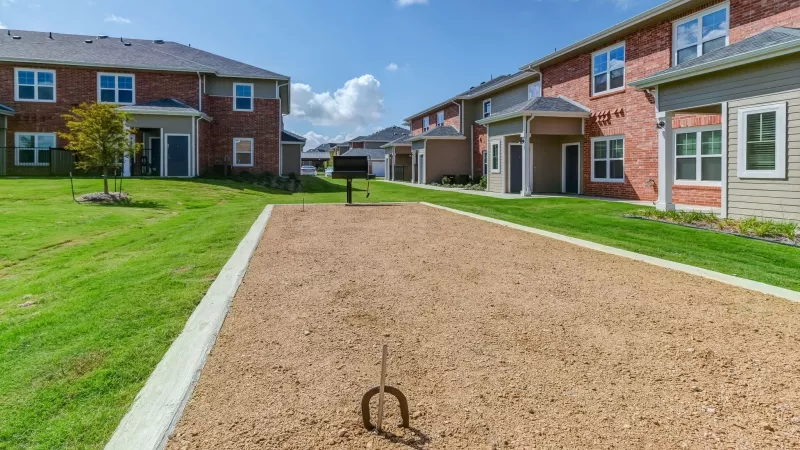 Horseshoe pit in a community lawn, flanked by multi-story brick apartment buildings under a blue sky.