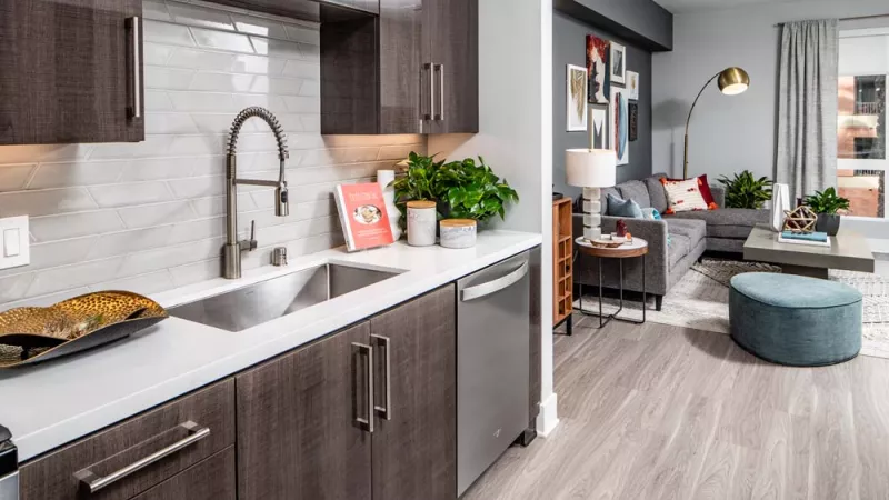 Modern kitchen with dark cabinets, white counters, and stainless steel sink. Open living room with grey sofa and teal ottoman.