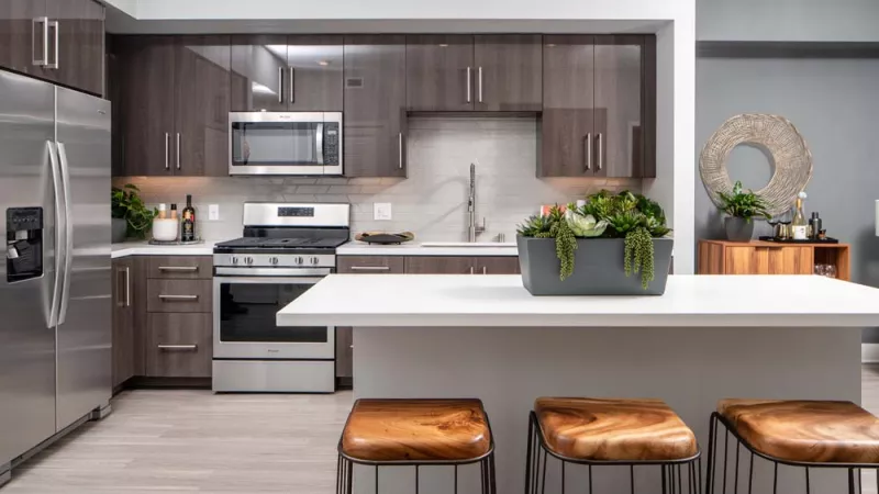 Modern kitchen with stainless steel appliances, dark cabinets, white island, three leather stools, and potted plants.