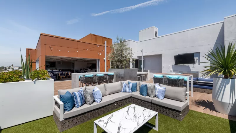 Modern rooftop patio with L-shaped sofa, marble coffee table, artificial grass, building views, and blue sky.