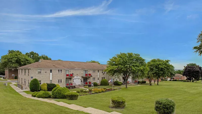 Panoramic view of a beige apartment building with red accents, lush green lawns, trees, and colorful flower beds under a blue sky.