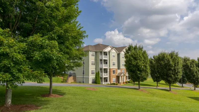 Photograph: Light green apartment building on a grassy hill, surrounded by trees under a blue, cloudy sky.