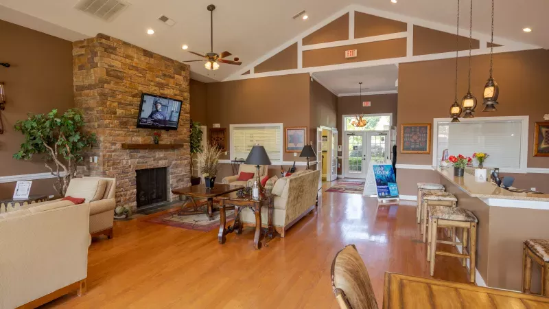Spacious community lounge with a stone fireplace, mounted TV, couches, wood floor, and a bright bar area.