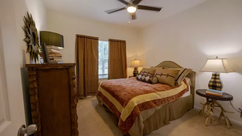 Warm bedroom with a patterned bedspread, wooden dresser, antler-leg bedside table, brown curtains, and a ceiling fan.