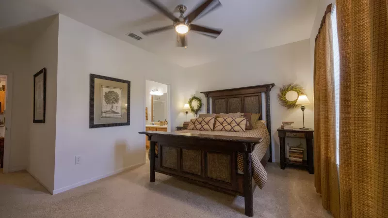 Bedroom with a large dark wood bed, ceiling fan, lamps on nightstands, and decorative wreaths. A glimpse of a bathroom.