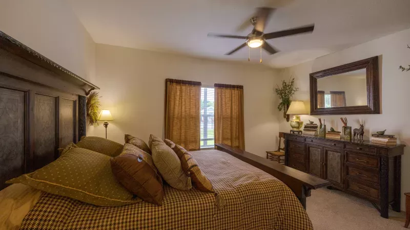 Cozy bedroom with a dark wood bed, golden and brown bedding. A matching dresser, mirror, and window are to the right.