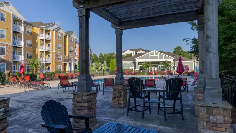 Sunny apartment complex pool deck with a pergola, red chairs, and a pool, surrounded by colorful buildings.