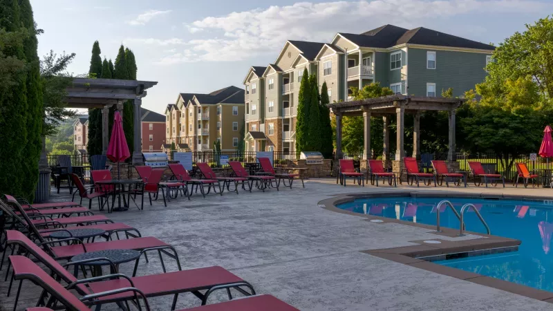 Outdoor swimming pool area with red lounge chairs, pergolas, and apartment buildings under a clear sky.