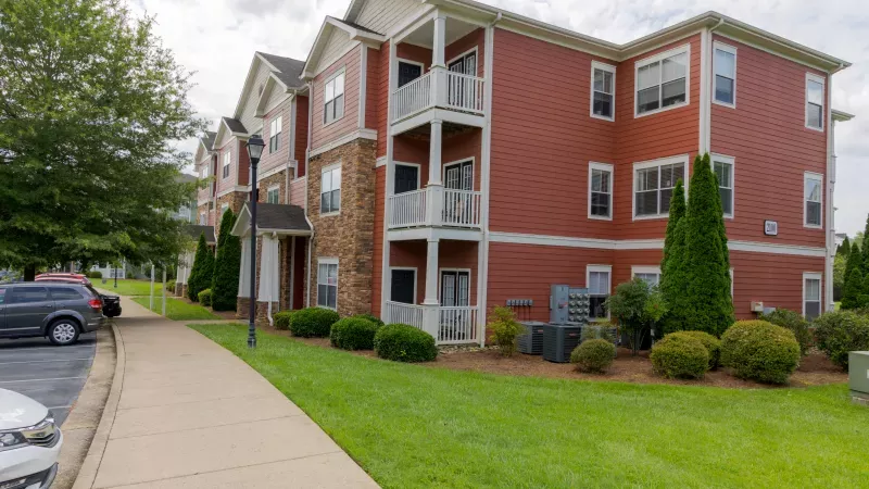 Red apartment building with stone accents, balconies, a sidewalk, green lawn, and parked cars under a cloudy sky.