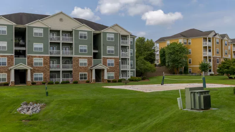 Photograph: Green and yellow apartment buildings overlook a grassy lawn with a sand volleyball court on a cloudy day.