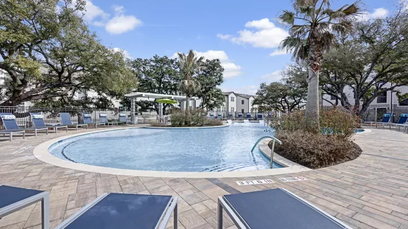 Spacious outdoor pool area with numerous blue lounge chairs, palm trees, and apartment buildings under a sunny sky.