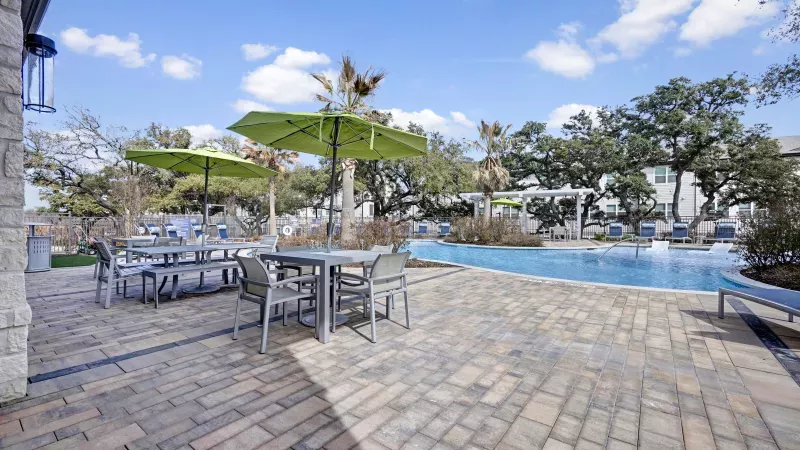 Outdoor patio with tables, green umbrellas, and a swimming pool under a blue sky.