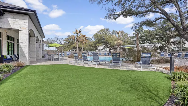 Outdoor pool area with a green lawn, lounge chairs, a blue pool, and a stone building, under a bright sky.