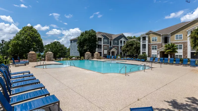 Outdoor swimming pool with blue lounge chairs, surrounded by apartment buildings and green trees under a sunny sky.