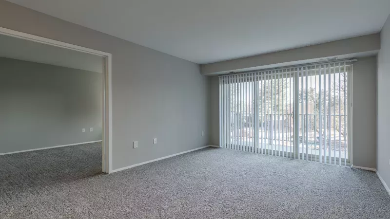 Empty living room with gray carpet and walls, a large sliding door with blinds opens to a balcony. Adjoining room visible.