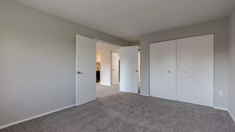 Unfurnished room with light gray walls, gray carpet, an open doorway to another room, and two closed closet doors.