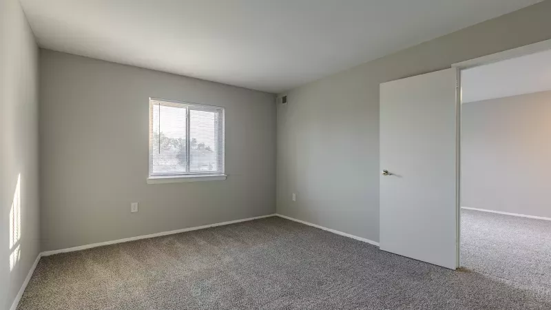 Empty bedroom with light gray walls, beige-brown carpet, and a window with white blinds. An open doorway leads to another room.