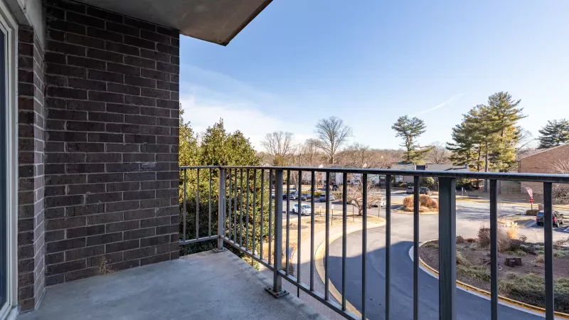 View from a balcony with a metal railing overlooking a curving road, trees, and buildings under a clear blue sky.