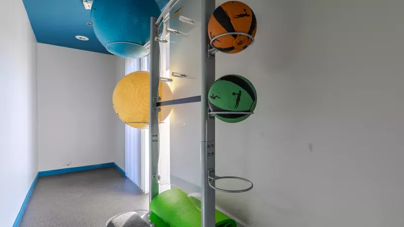 Bright fitness room with a metal rack holding colorful exercise balls. A green yoga mat is on the floor.