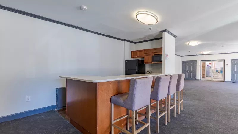 Community lounge with a wooden bar, four purple stools, and a small kitchen area with a microwave.