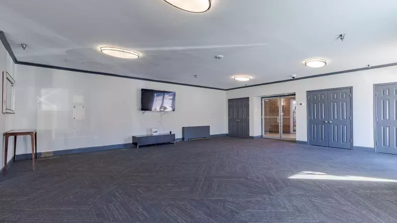 Large empty room with geometric blue carpet, white walls, mounted TV, and multiple dark grey doors.