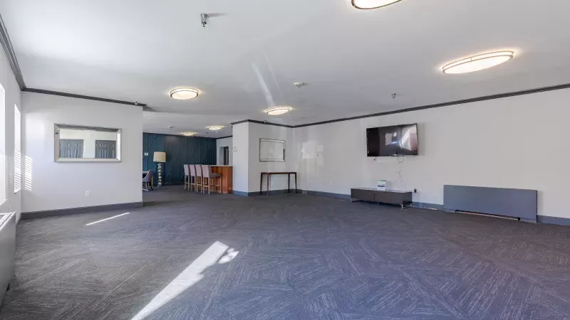 Empty common room with dark blue patterned carpet, light walls, wall-mounted TV, and bright ceiling lights.